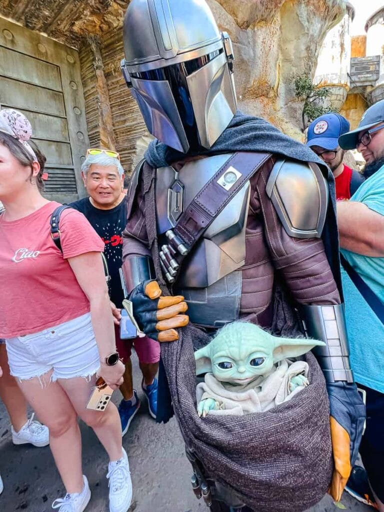 "The Mandalorian wearing armor holding Baby Yoda (Grogu) in a desert landscape"