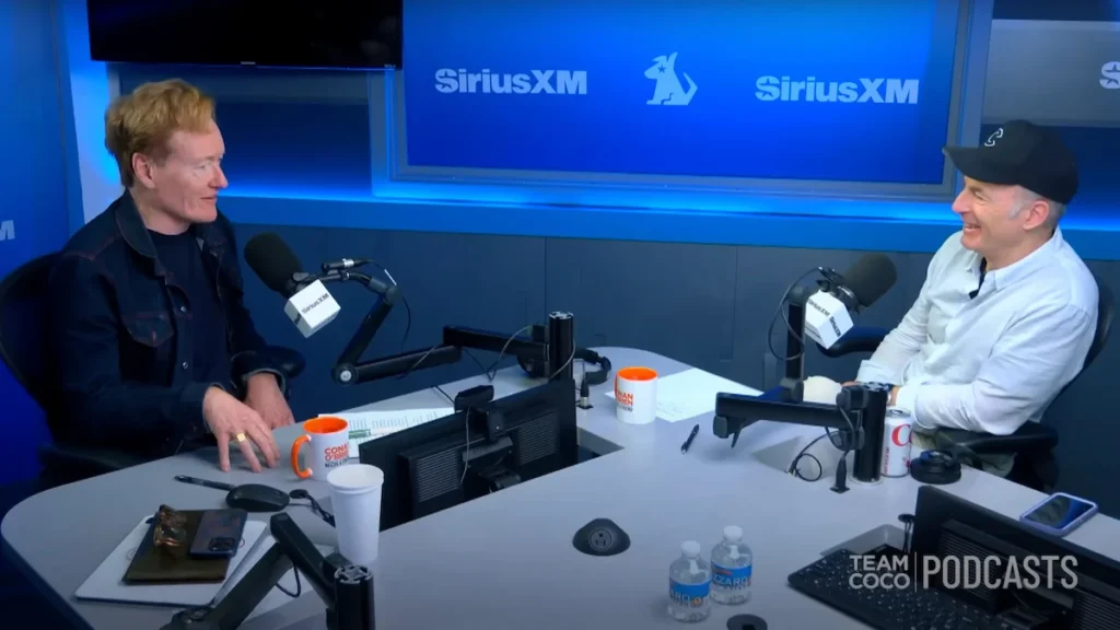 “Conan O’Brien wearing headphones and smiling while recording his comedy podcast.”