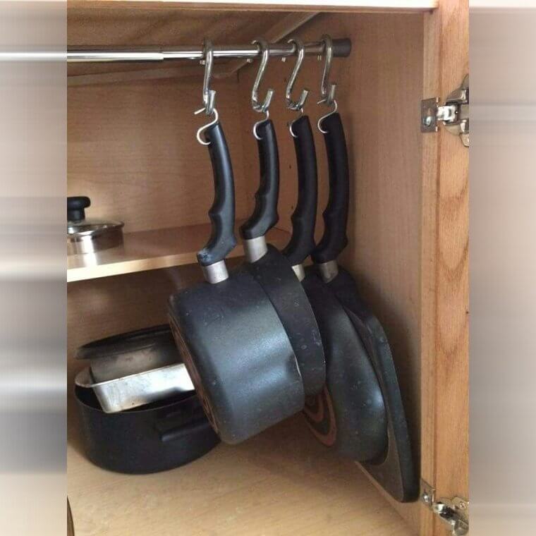A kitchen wall or cabinet with pots and pans hanging from a tension rod using S-hooks for easy access and display.