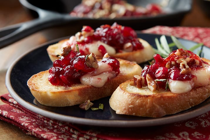 “Warm baked brie topped with cranberry sauce and pecans on a holiday serving board.”