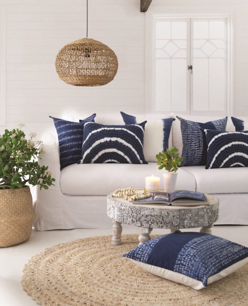 Coastal living room with a woven rattan pendant light, indigo pillows, carved wood coffee table, and jute rug.