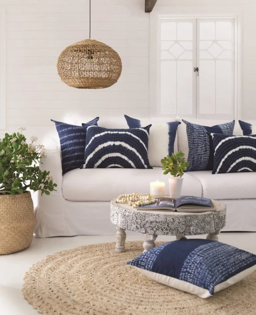 Coastal living room with a woven rattan pendant light, indigo pillows, carved wood coffee table, and jute rug.