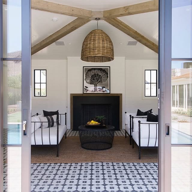 Indoor-outdoor living space with a bold rattan pendant light, vaulted wooden beams, black fireplace, and patterned tile flooring.