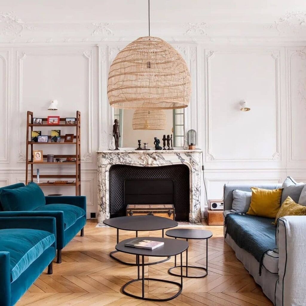 Luxe living room featuring an oversized rattan pendant light, velvet chairs, marble fireplace, and elegant decorative accents.
