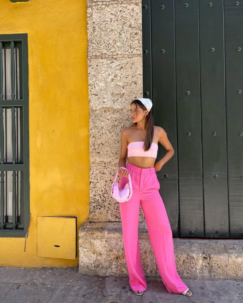 A pastel pink bandeau top with matching flowy trousers, styled with a braided pink handbag, white hair scarf, and open-toe sandals.