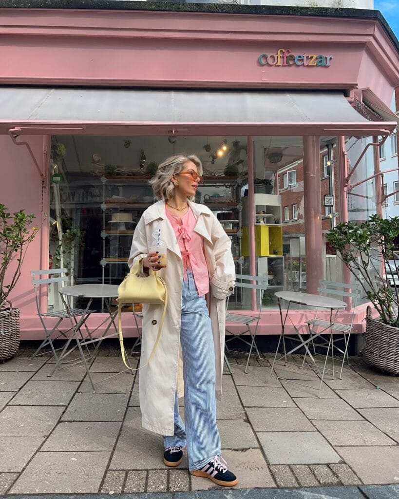 Light pink bow-detail blouse paired with striped wide-leg jeans, trench coat, sneakers, and a pastel yellow handbag.