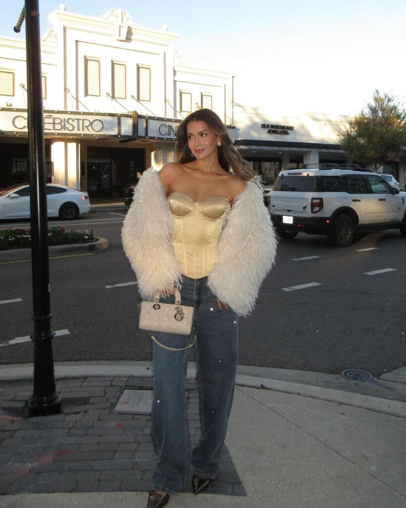 A gold structured corset top paired with sparkly wide-leg jeans, a fluffy white jacket, metallic heels, and a designer handbag.