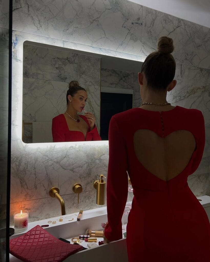 A red fitted dress with long sleeves and a heart-shaped cutout in the back, styled with a pearl necklace, updo hairstyle, and red clutch.
