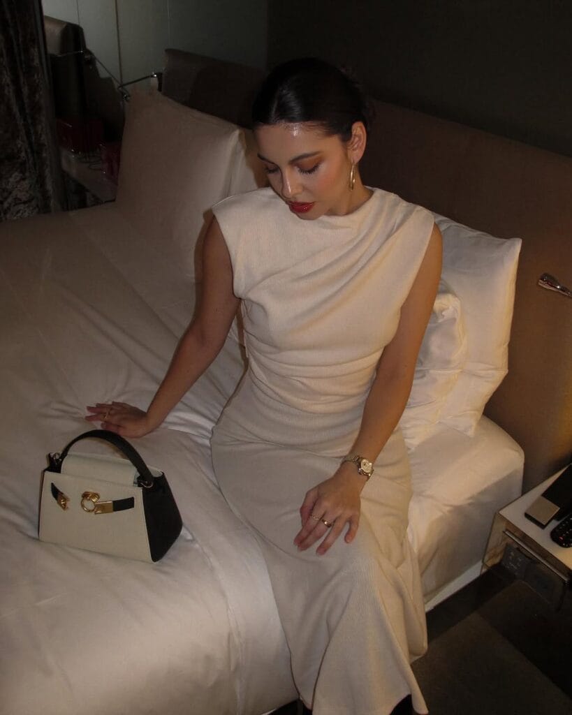 Cream midi dress with soft draping, styled with a black-and-white structured handbag, gold jewelry, and red lipstick.
